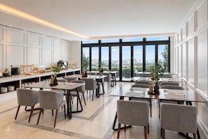 Un área de comedor moderna con varias mesas y sillas, una barra de cocina con electrodomésticos y amplios ventanales con vistas a la ciudad.