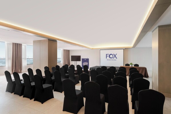 A conference room with rows of black chairs, a stage with a podium, and a screen displaying "FOX" and "Glenmarie Shah Alum".