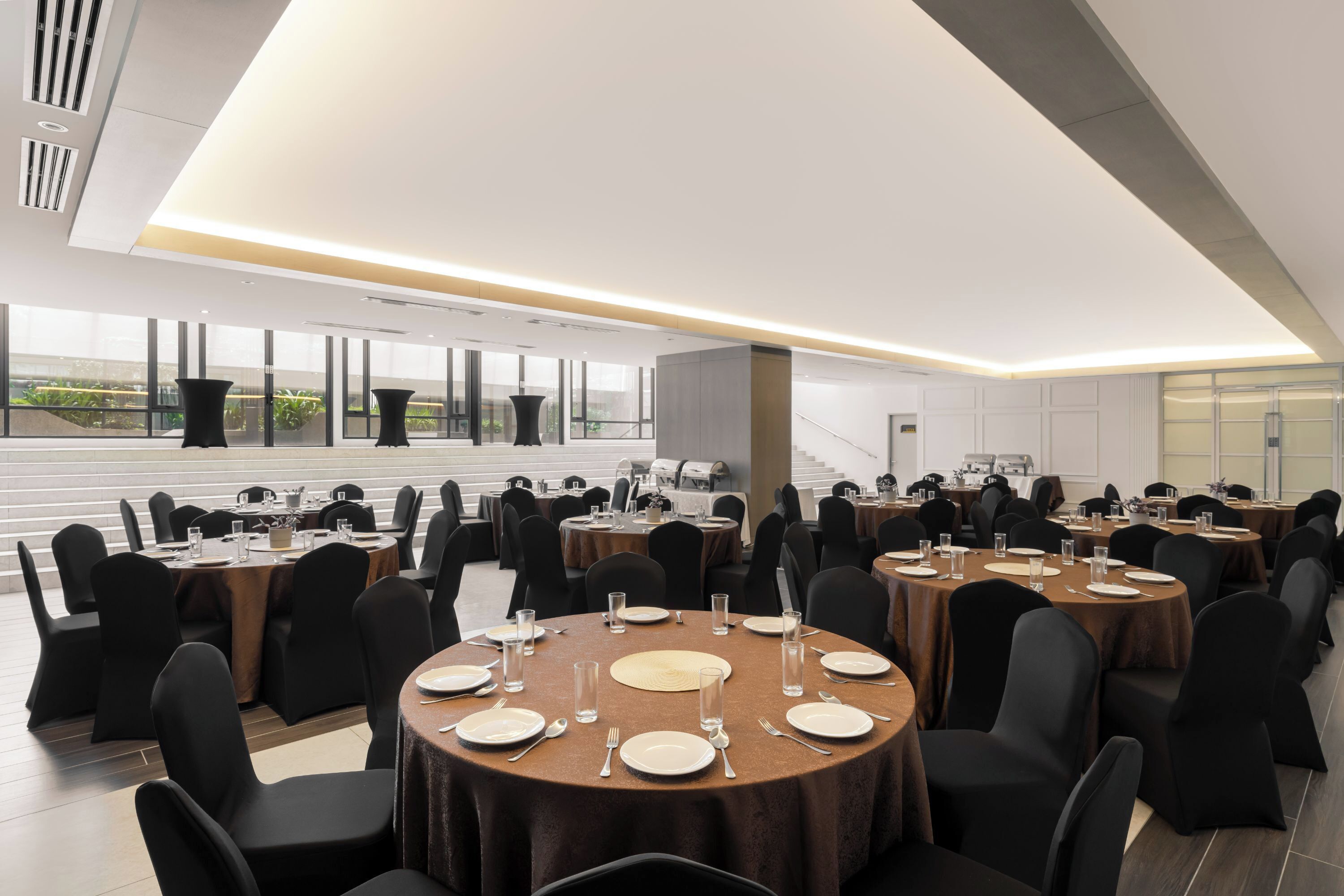 A spacious conference room with multiple round tables, black chairs, and a large window offering a view of greenery.
