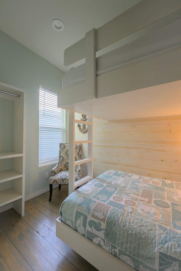 A bunk bed with a quilt, a chair, and a shelving unit in a room with wooden flooring and a window with blinds.