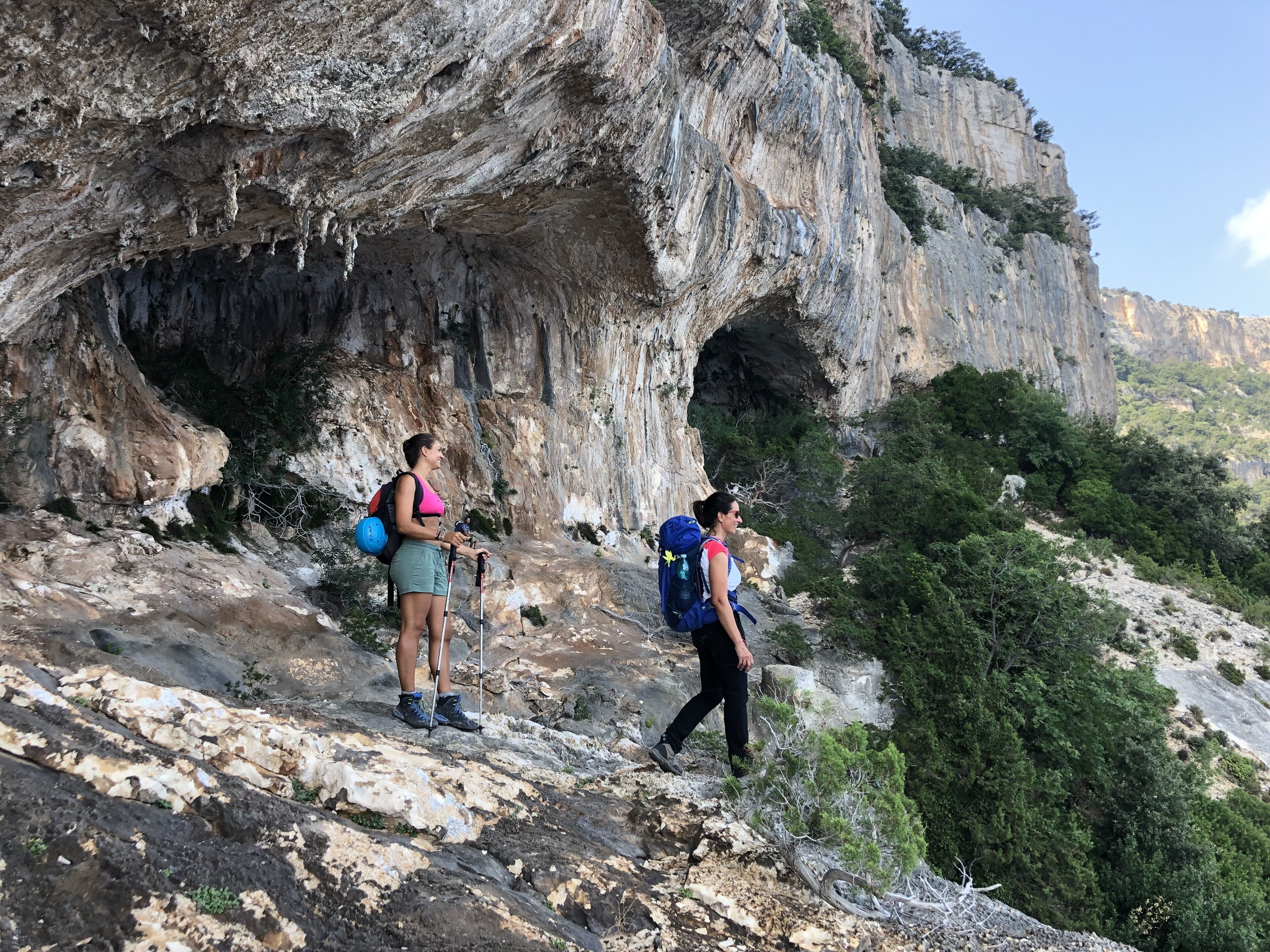 Two hikers on a rocky path with a large cave-like rock formation in the background.
