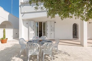Un bâtiment blanc doté d’un patio où se trouve une table dressée pour le repas, entourée de chaises blanches.