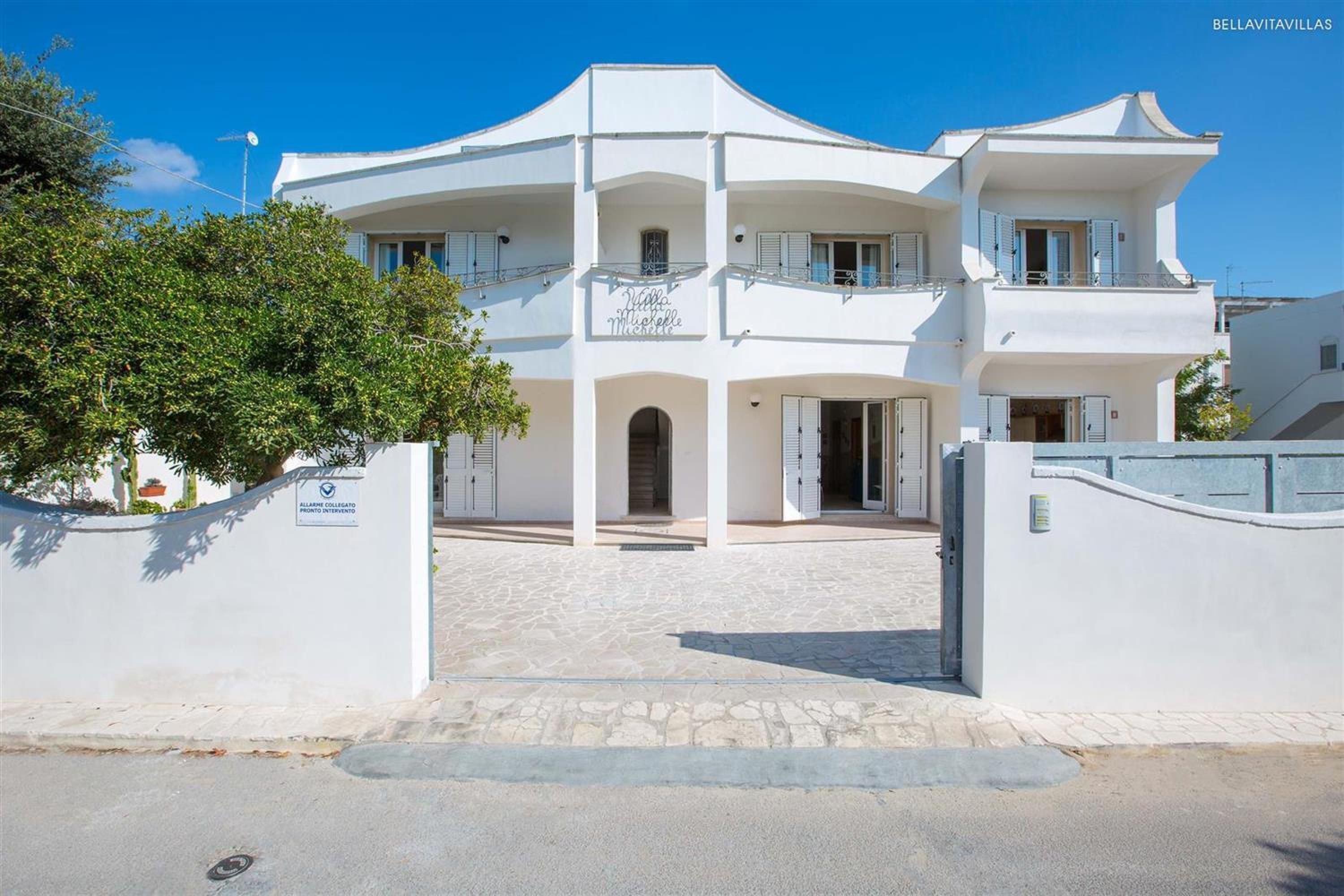 Un edificio blanco de dos pisos con balcones, un letrero que dice "Villa Michele" y un área de entrada pavimentada.