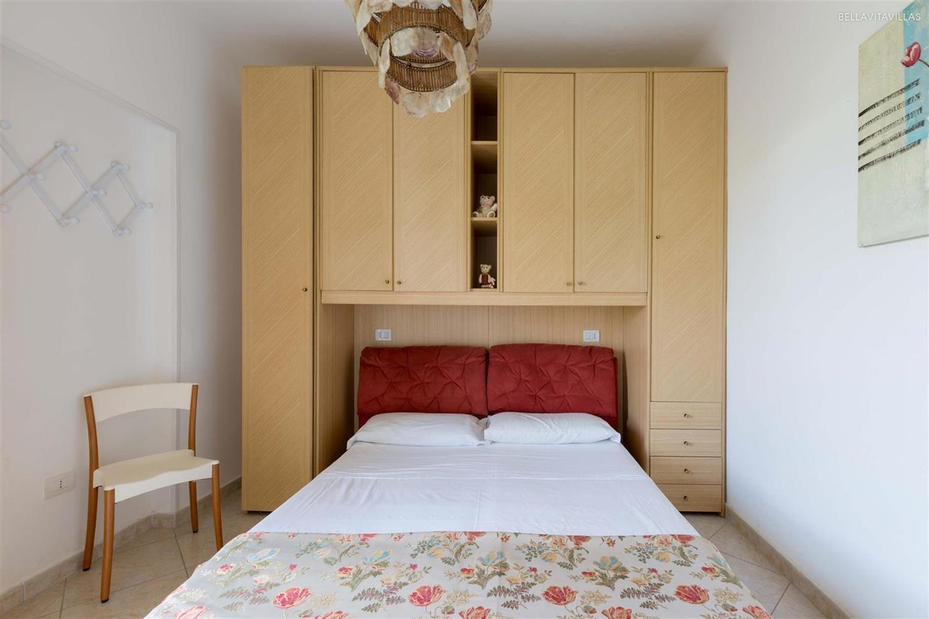 A bedroom with a bed, a wooden wardrobe, a chair, and a wall-mounted light fixture.