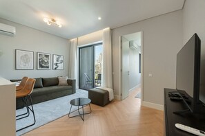 Un salon moderne avec un canapé, des fauteuils, une table basse et un téléviseur. Il y a un balcon avec un fauteuil et une rambarde.