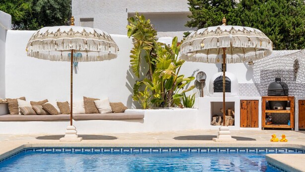 Un espace piscine agrémenté de deux parasols blancs, d’un canapé avec des coussins et d’un barbecue.