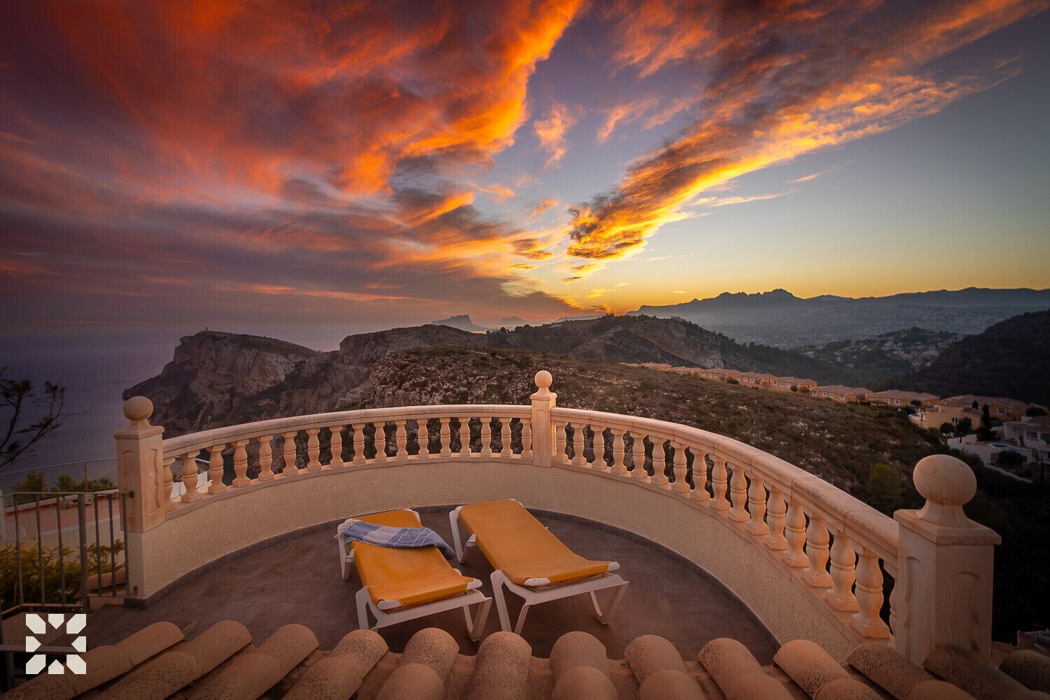 Un balcon doté de deux chaises longues, d’une balustrade en pierre et offrant une vue sur un paysage côtier avec des montagnes et un coucher de soleil.