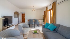 Un soggiorno moderno con un divano grigio, due poltrone, un tavolino da caffè con una mela verde, una TV su un mobile in legno e un'area pranzo con tavolo e sedie.