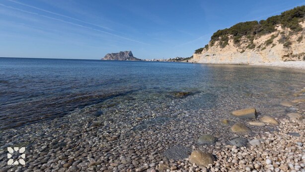 Ein Kiesstrand mit klarem Wasser, einem Felsen und einer entfernten Stadtansicht.