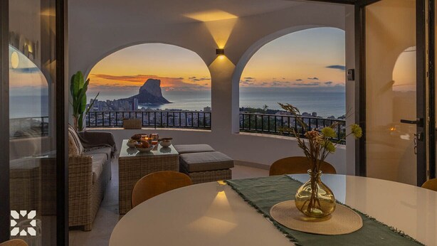 Une chambre moderne avec un balcon donnant sur un paysage urbain et l’océan au coucher du soleil.