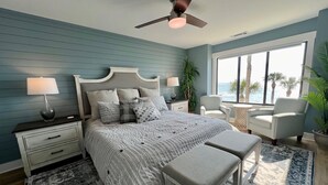 Une chambre à coucher avec un lit, des tables de chevet, un ventilateur de plafond, une fenêtre donnant sur un beau paysage et une plante en pot.