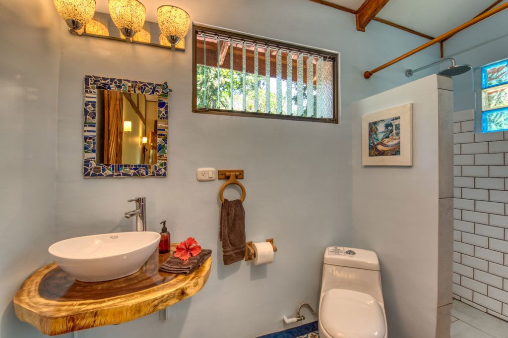 Un baño con tocador de madera, lavamanos blanco, inodoro, espejo con marco de mosaicos y ventana con vistas a la vegetación.