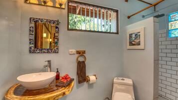 Un baño con tocador de madera, lavamanos blanco, inodoro, espejo con marco de mosaicos y ventana con vistas a la vegetación.