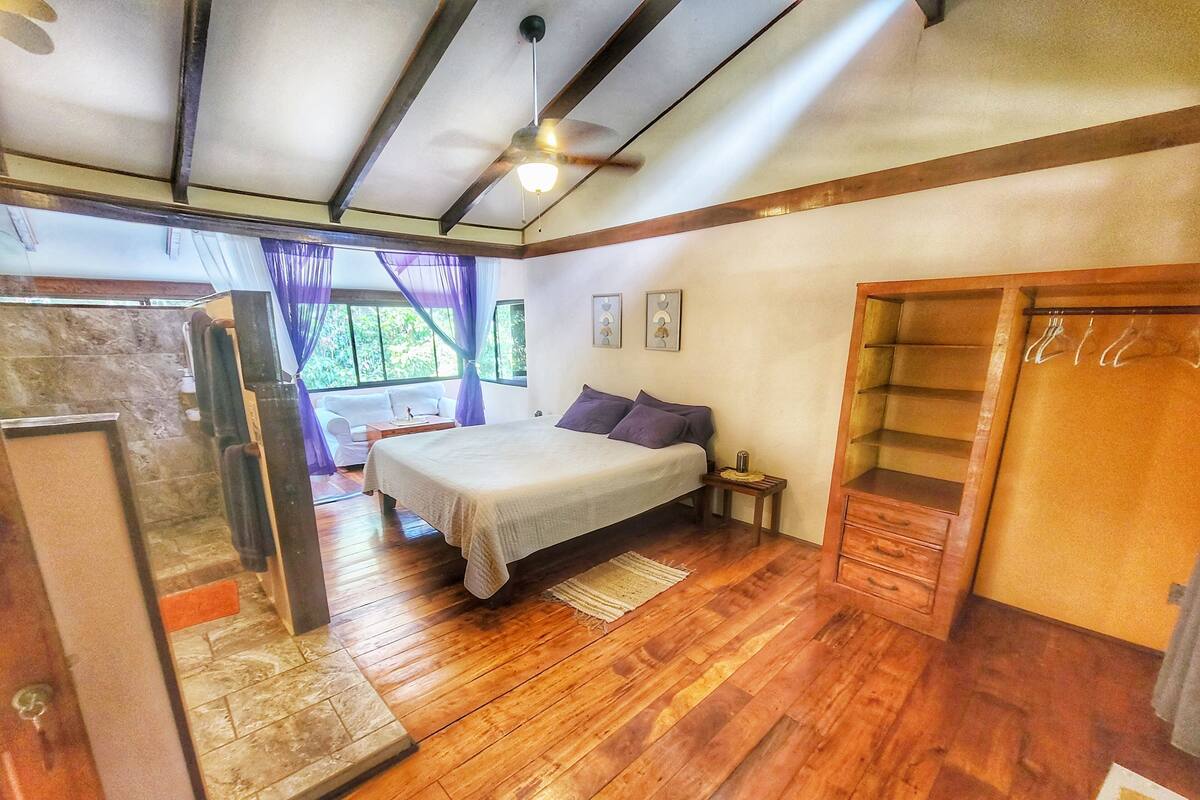 Un dormitorio con cama, suelo de madera, ventilador de techo, ventana con cortinas y armario.