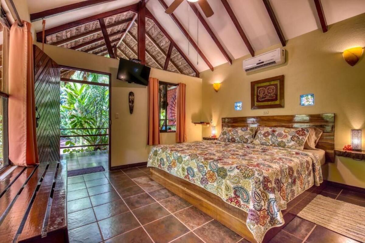 Un dormitorio con cama, televisor, equipo de aire acondicionado y vista a la vegetación por una puerta.