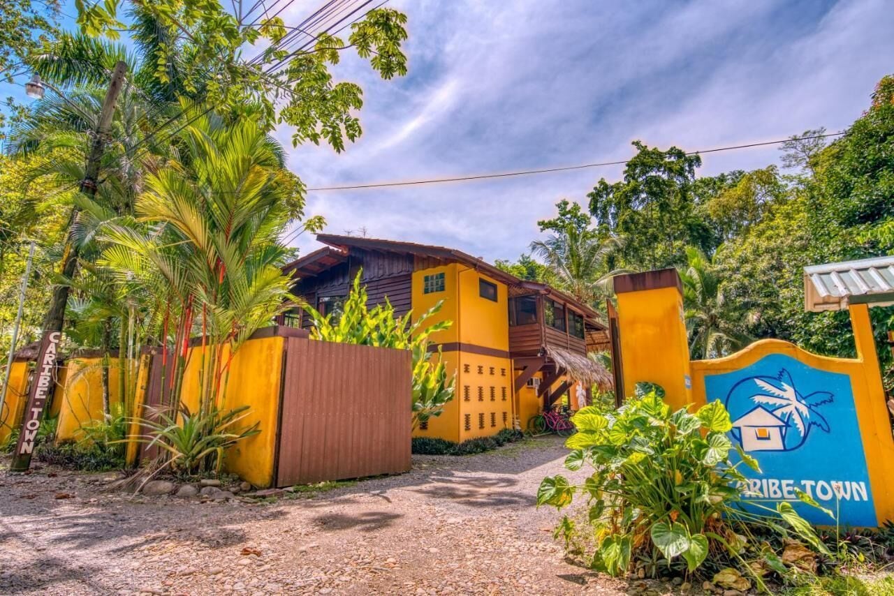 A rustic house with a yellow facade, surrounded by lush greenery and a gravel path leading to a wooden gate with a sign reading "CARIBE-TOWN".