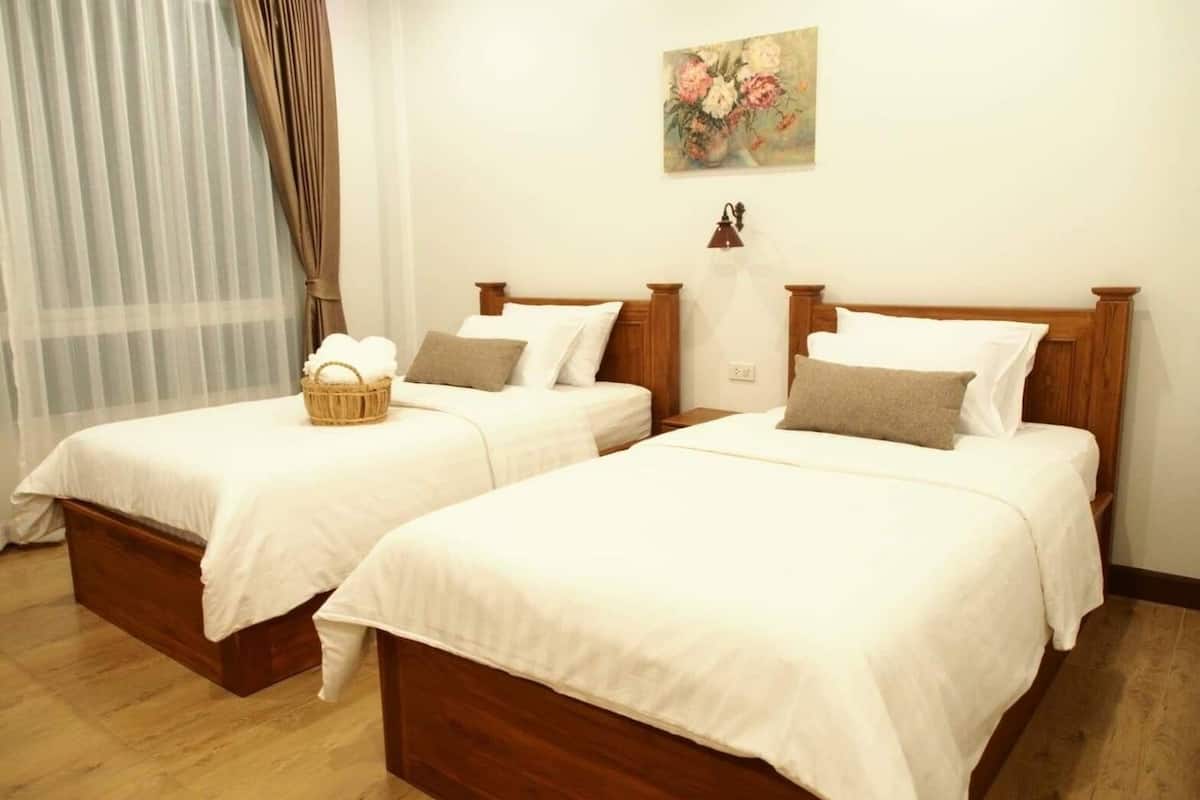 A hotel room with two wooden beds, white bedding, a wicker basket on one bed, and a floral painting on the wall.