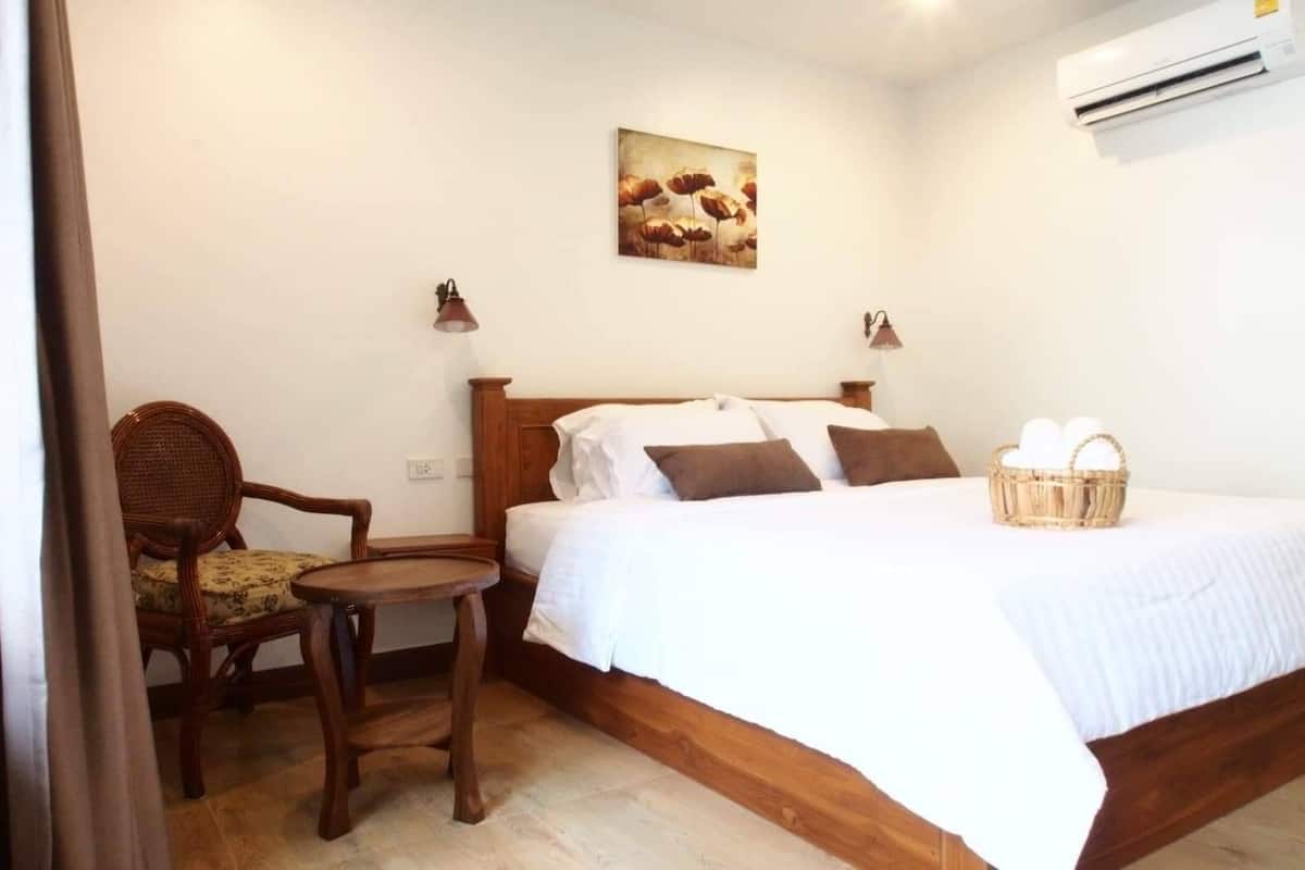 A bedroom with a wooden bed, a chair, a small table, a wall-mounted lamp, and a basket of eggs.