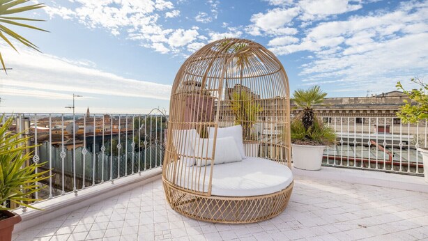 A round, wicker-framed outdoor seating area with white cushions on a rooftop terrace with a city view. - ARIA - Penthouse & Garden (Roma)