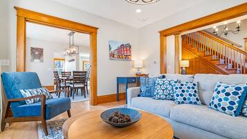 A living room with a blue armchair, a grey sofa with patterned cushions, a wooden coffee table, and a staircase leading to an upper floor.