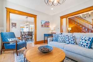 Un salon avec un fauteuil bleu, un canapé gris orné de coussins à motifs, une table basse en bois et un escalier menant à l’étage supérieur.
