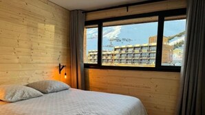 Ein Schlafzimmer mit einer Holzwand, einem Bett mit grauer Bettdecke, einem Fenster mit Vorhängen und Blick auf Gebäude und Berge.