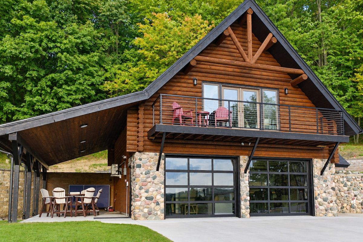 Eine Blockhütte mit Steinfundament, einer großen Garagentür und einer überdachten Terrasse mit Sitzgelegenheit im Freien.