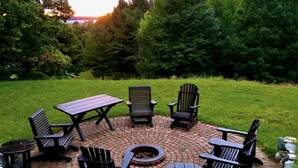 Un espacio de estar al aire libre circular con una hoguera, rodeado de sillas tipo Adirondack y una mesa de picnic.