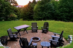Et cirkulært udendørs opholdsområde med bålfyr, omgivet af Adirondack-stole og et picnicbord.