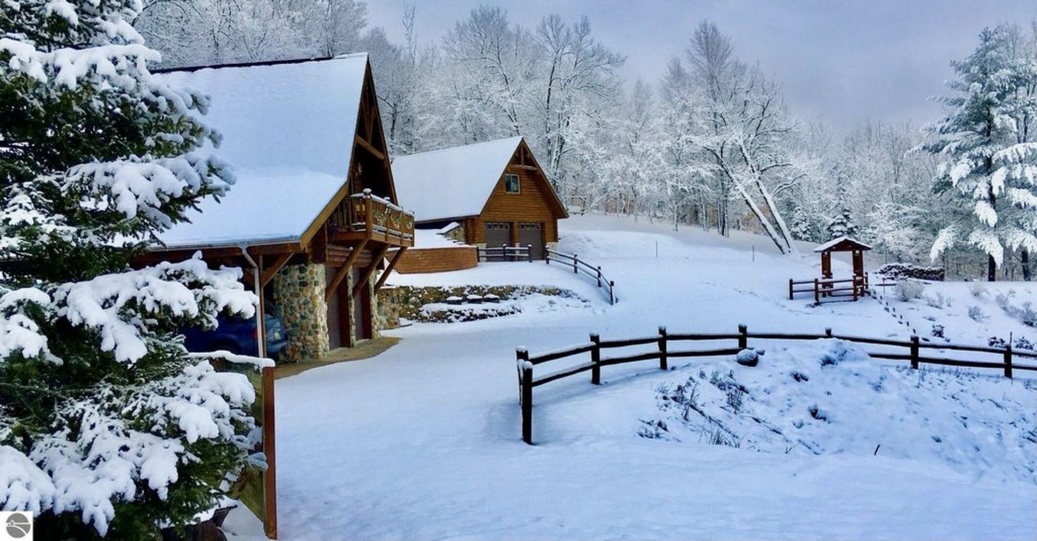 A wooden cabin with a snow-covered roof, surrounded by a wooden fence and snow-covered trees.