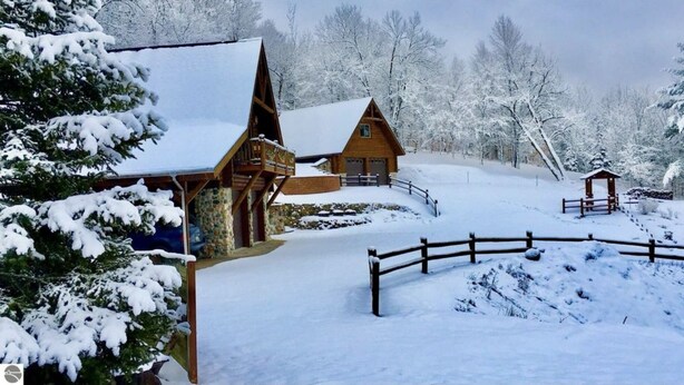 Una cabaña de madera con techo nevado, rodeada por una valla de madera y árboles cubiertos de nieve.
