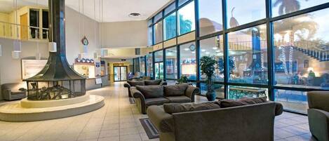 A spacious lobby with a large fireplace, comfortable seating, and large windows offering views of palm trees and a building outside.