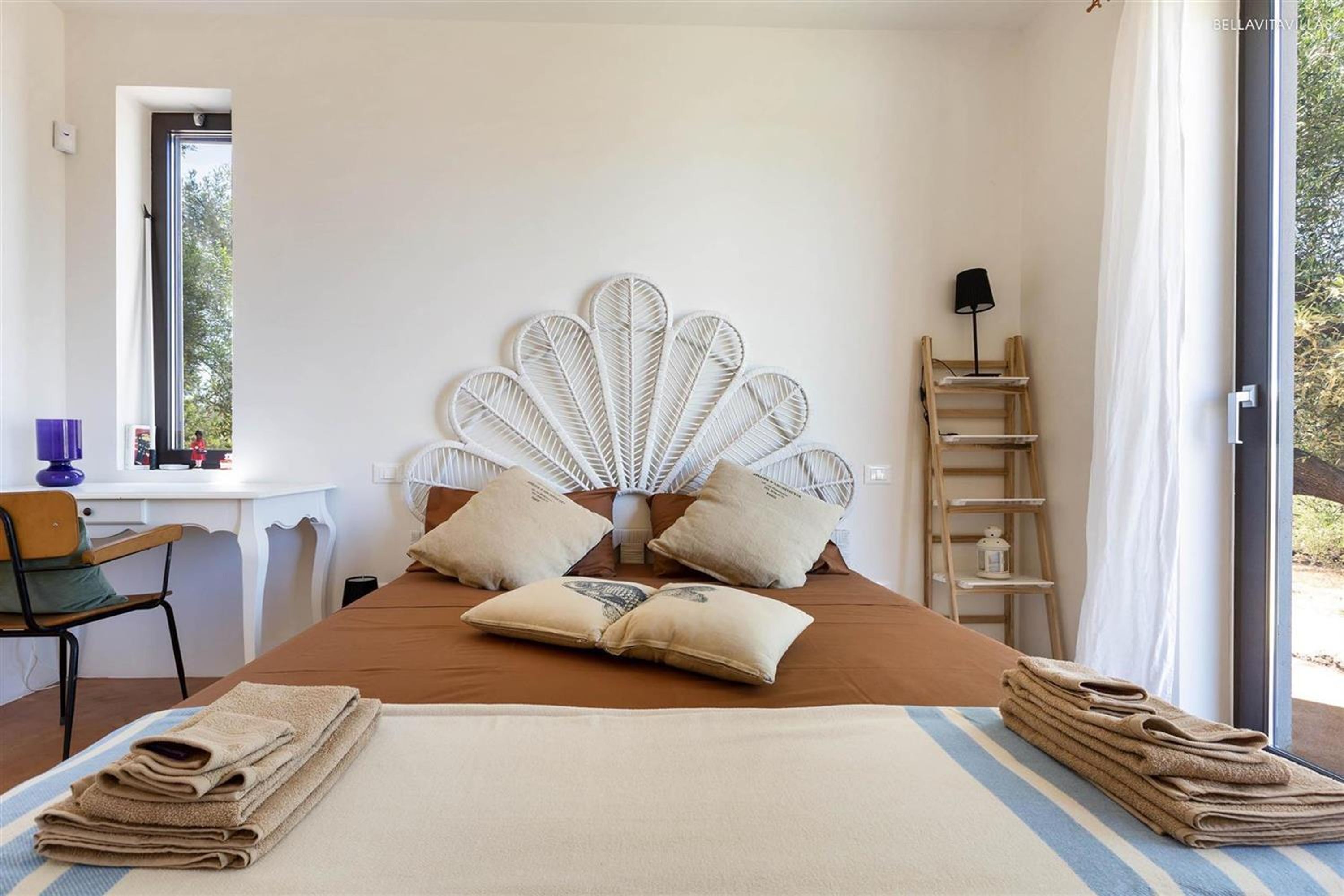 A bedroom with a bed, pillows, a bedside table, a chair, a window, and a view of greenery outside.