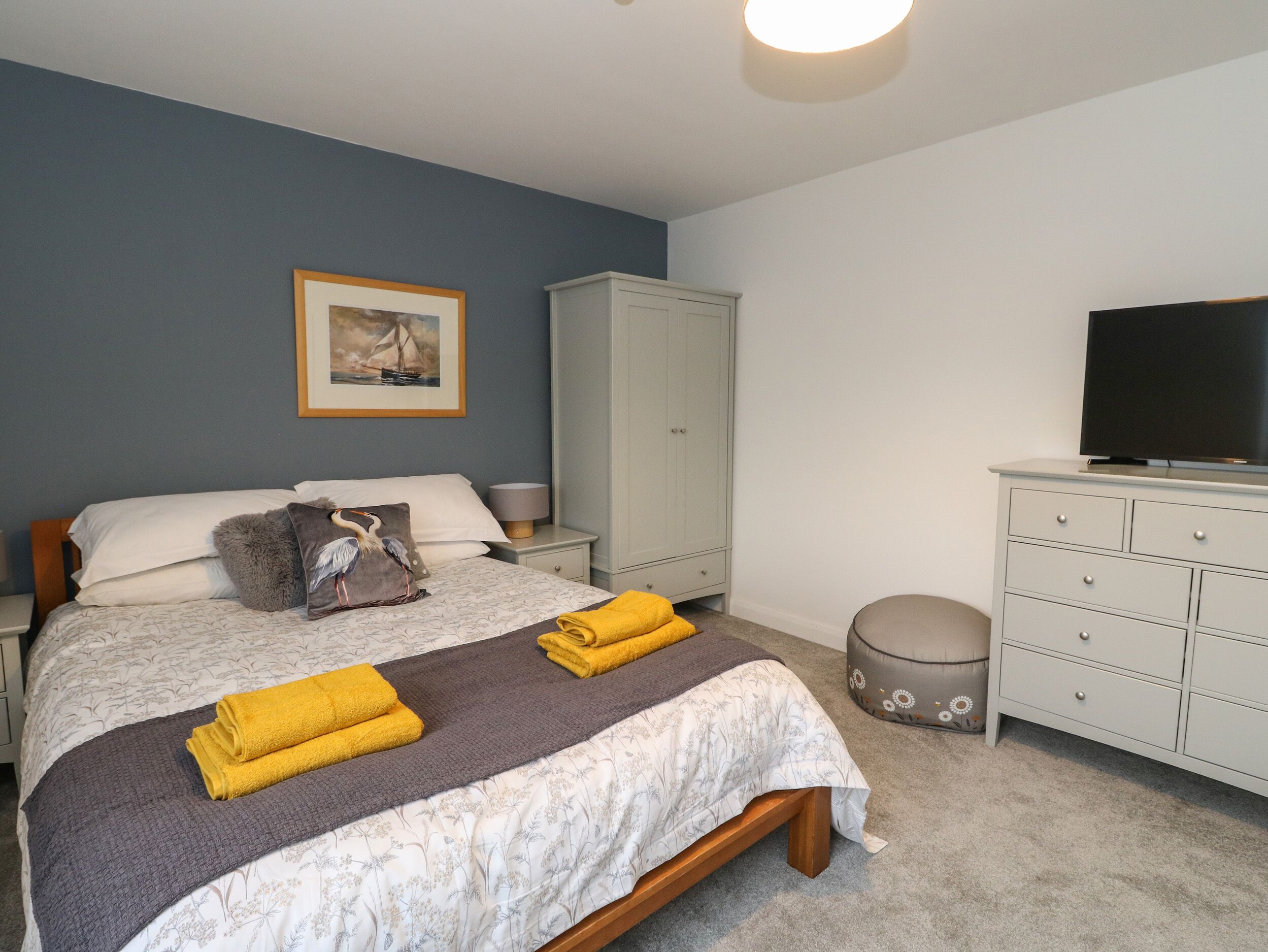 A neatly made bed with a floral bedspread and two folded yellow towels, a white wardrobe, a television on a stand, and a framed picture on the wall.