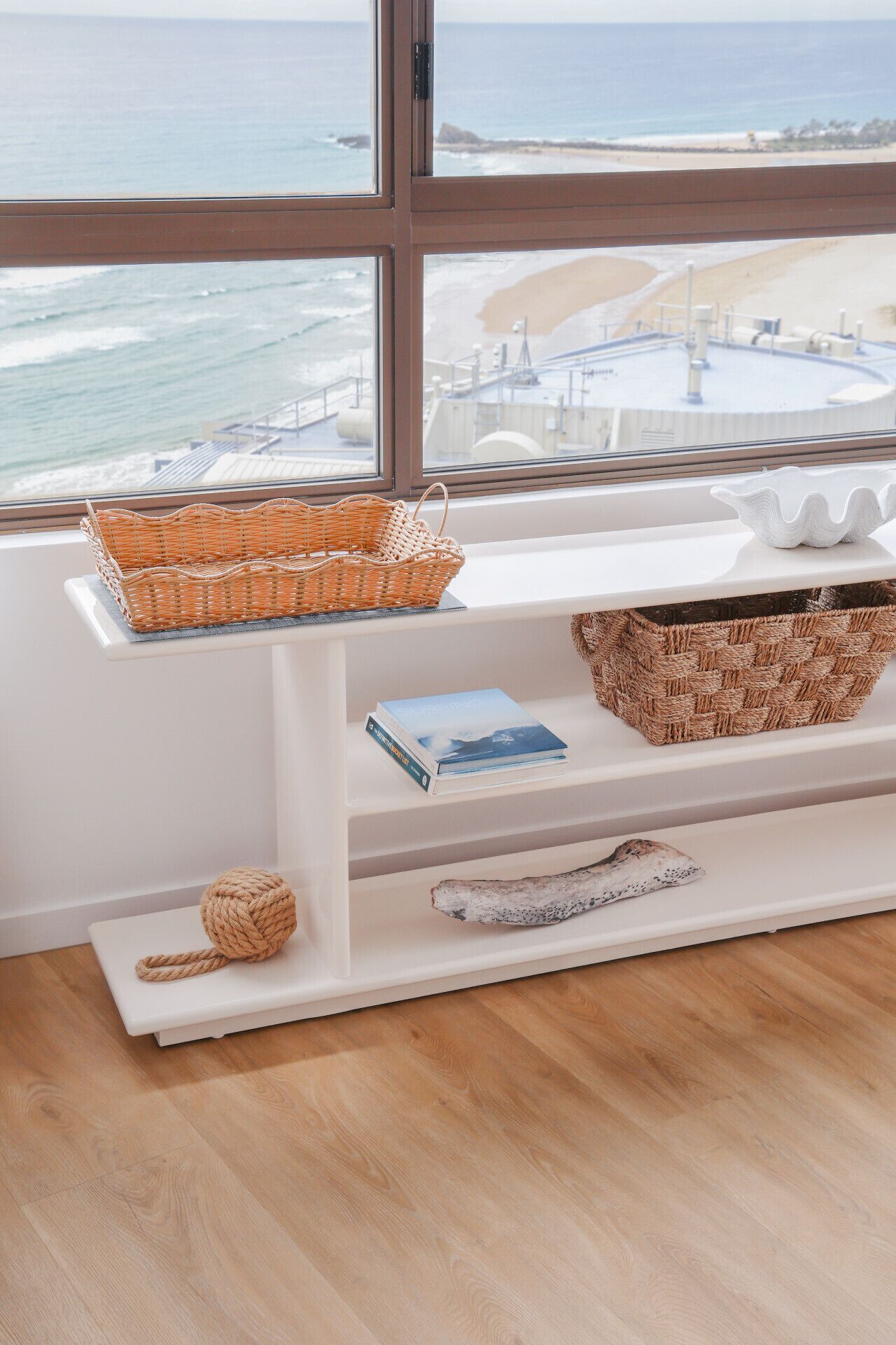 A room with a large window overlooking a beach and ocean. There is a white shelf with a woven basket, a book, and a decorative item.