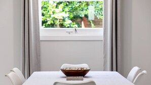 Et spisebord med en skål og en mindre genstand, et vindue med udsigt til grønne omgivelser, og hvide stole omkring bordet.