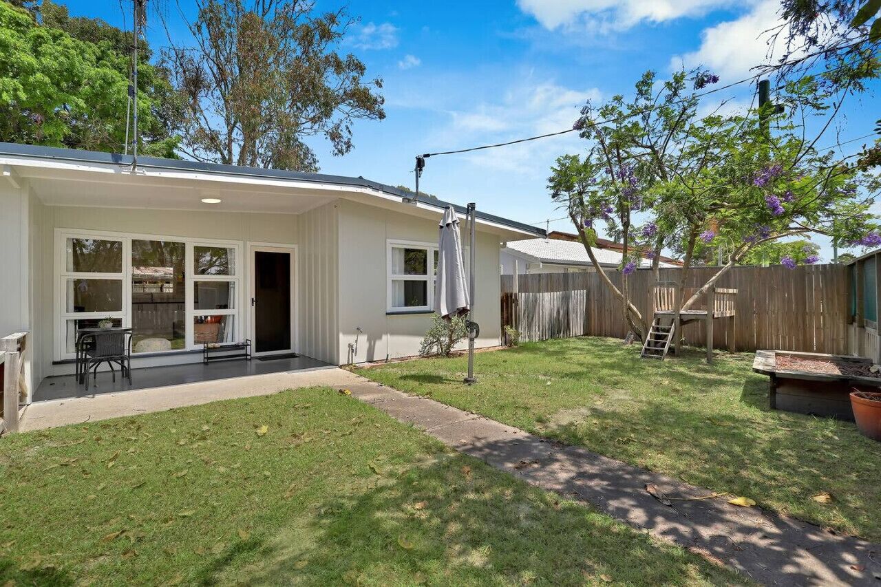 A single-story house with a covered patio, a fenced backyard, and a tree with purple flowers.