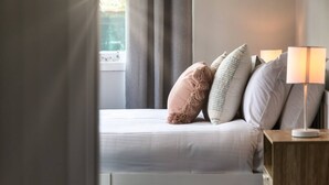 En pænt redt seng med et hvidt dynebetræk, et trænatbord med en lampe, og et vindue med gardiner.