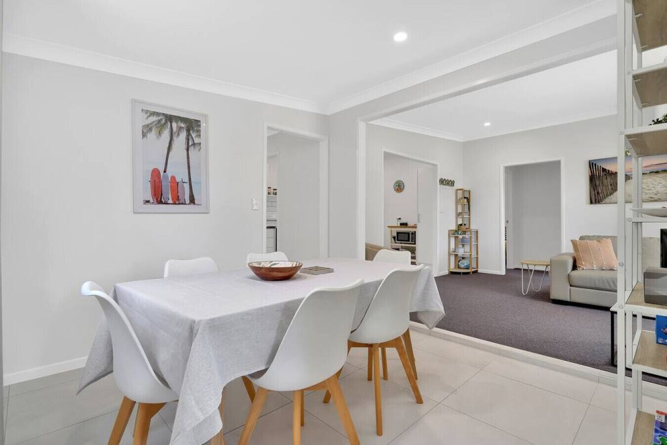 Un comedor con una mesa preparada para dos, un tazón sobre la mesa y una silla blanca. Se ve una estantería con libros y una pequeña mesa con una silla. Al fondo, un sofá y un televisor.