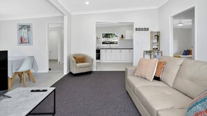 En moderne stue med en beige sofa, en mindre lænestol og et fjernsyn på et marmorbord. Et køkken er synligt i baggrunden med hvide skabe og et vindue.