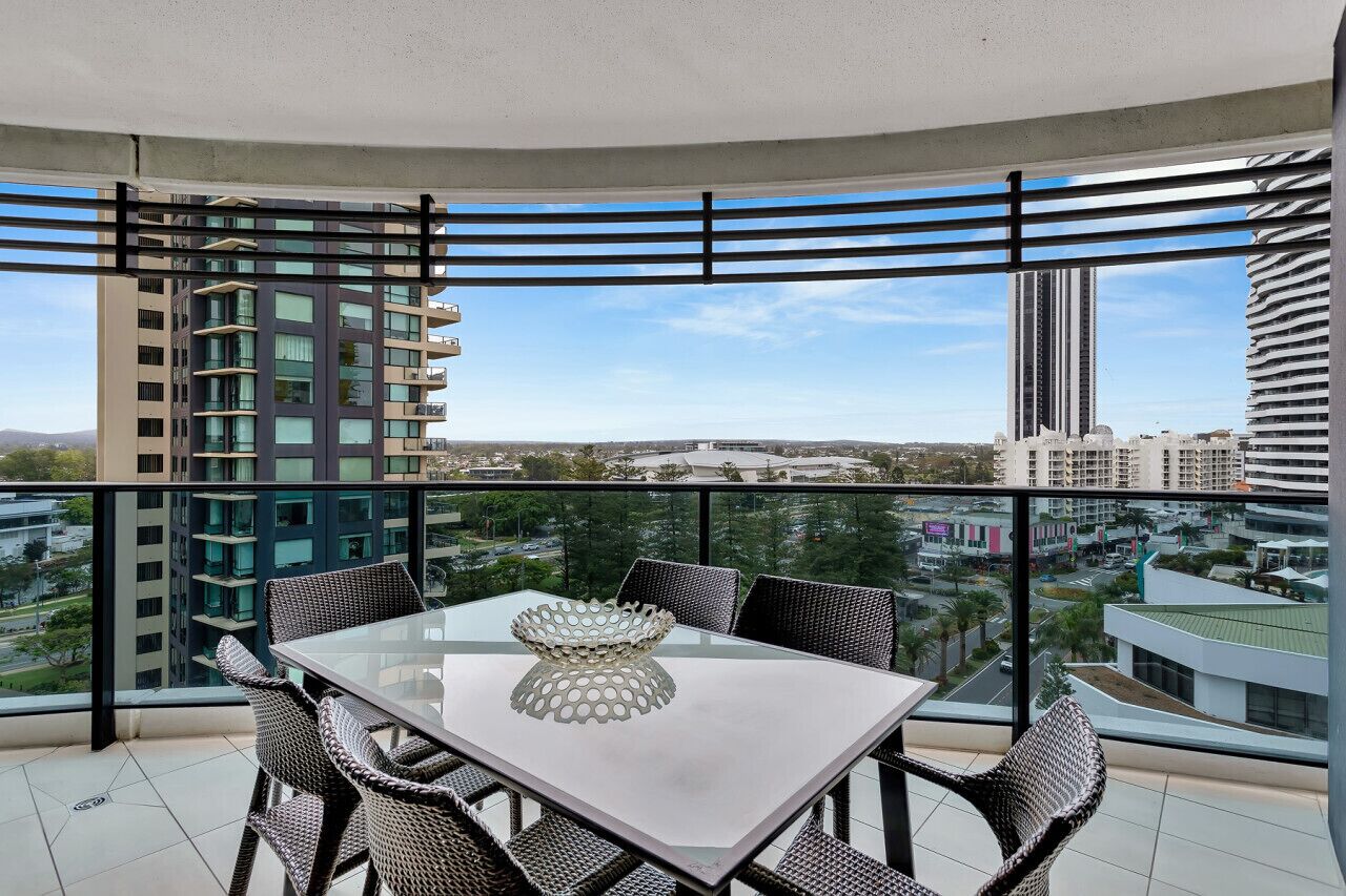 Un balcon doté d’une table en verre et de chaises en osier, offrant une vue sur un paysage urbain avec des bâtiments modernes.