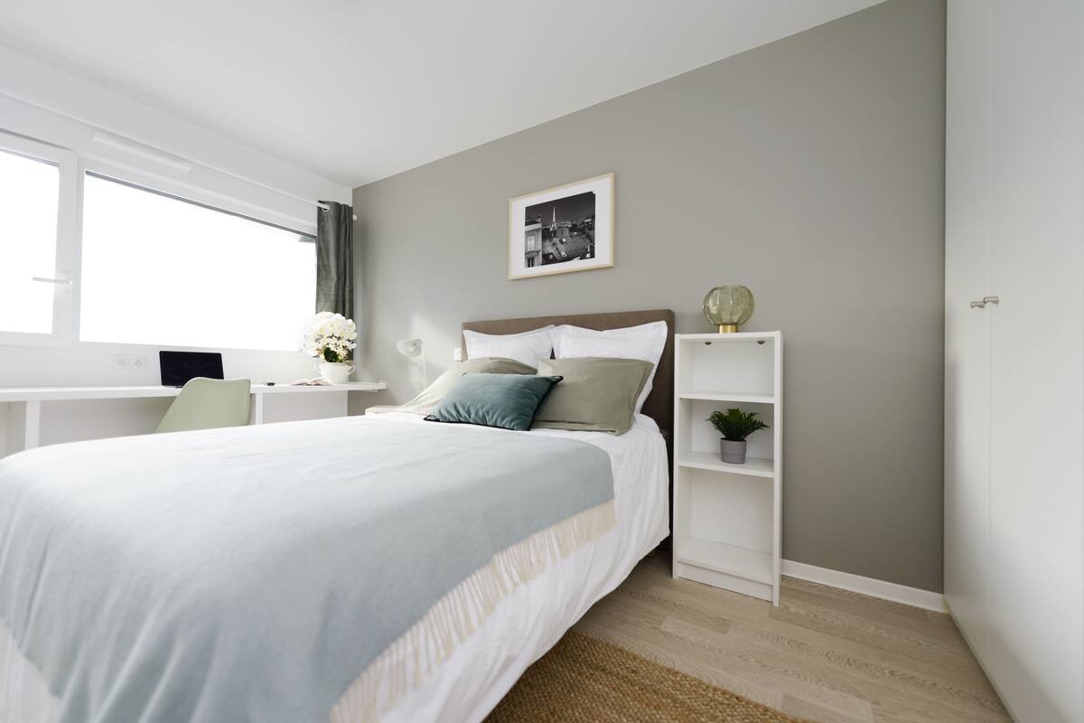 A bedroom with a bed, a desk, a chair, a shelf with a plant, and a framed picture on the wall.