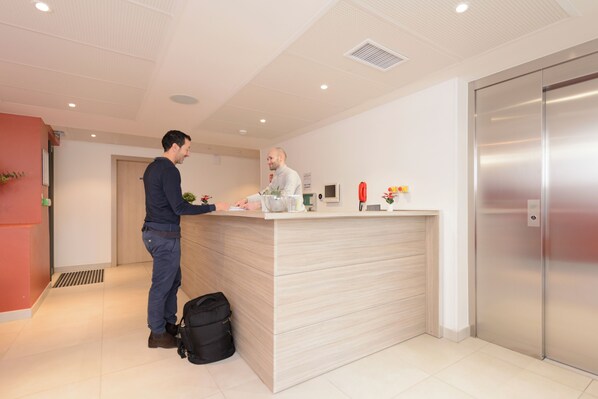 A reception desk with a man standing at it, a black bag on the floor, and an elevator to the right.