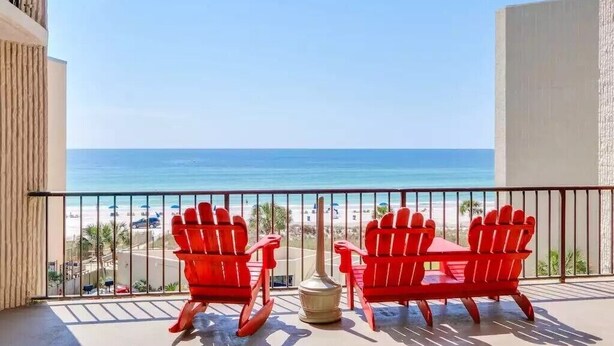 Ein Balkon mit zwei roten Adirondack-Stühlen und einem kleinen Tisch mit Blick auf einen Strand und einen klaren Meerblick.