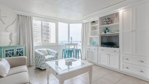 Ein Wohnzimmer mit einem weißen Sofa, einem Glastisch, einem Fernseher auf einer weißen Kommode und Meerblick durch große Fenster.