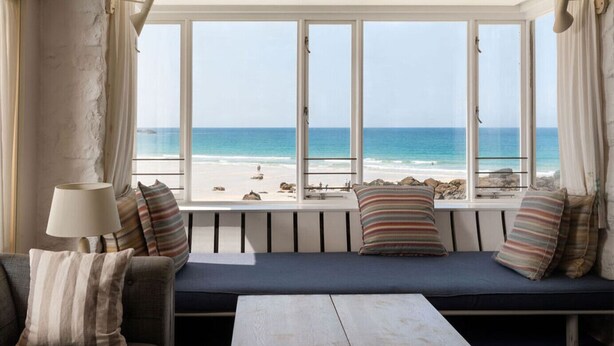 A room with a large window overlooking a beach, a sofa with striped cushions, and a lamp on a side table. - Old Saltings, overlooking Porthmeor beach (St Ives)