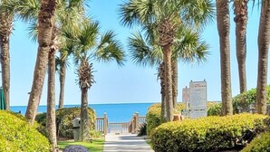 A pathway lined with palm trees leading to a beach, with a stone sculpture in the foreground. - Romantic 14th floor w/ a WOW view! Oceanfront King, full condo, pools & hot tubs (Myrtle Beach)
