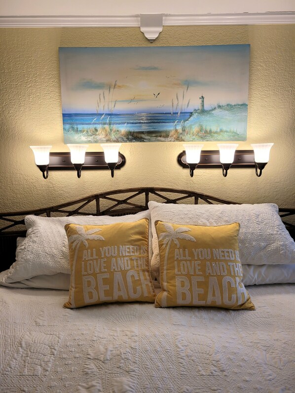 A bedroom with a bed, two wall-mounted light fixtures, a painting of a beach scene, and two pillows with text.