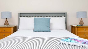 Un lit bien fait, recouvert d’une couvre-lit à rayures, avec un seul oreiller bleu et deux tables de chevet sur lesquelles sont posées des lampes et des livres.