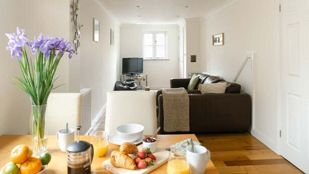 Un coin repas avec une table mise pour le petit-déjeuner, comprenant des croissants, des fruits et du jus d’orange. Un vase rempli de fleurs est posé sur la table, et on aperçoit un salon avec un canapé et un téléviseur au fond.
